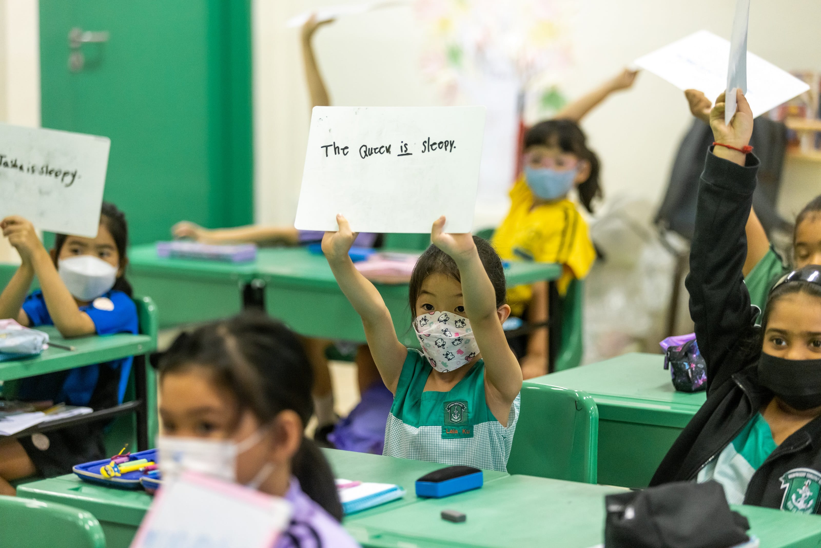 Young masked students sit at green desks holding up whiteboards with sentences like "The Queen is sleepy."